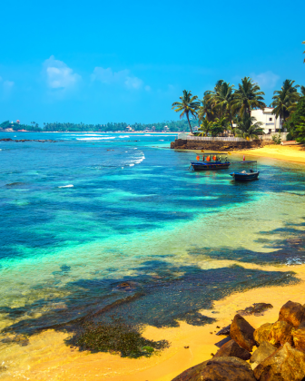 Turquoise shallows and golden sand at Mirissa beach, fishing boats anchored near palm-fringed shore.